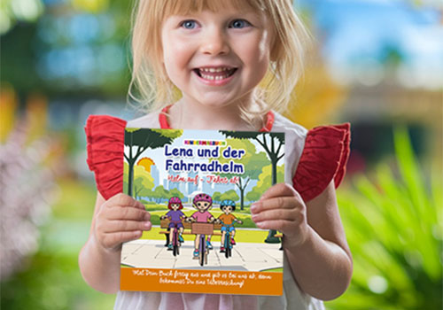 Lena und der Fahrradhelm - Kindermalheft für das Krankenhaus. Blondes Mädchen hält lächelnd das Malheft ‚Lena und der Fahrradhelm – Helm auf, Fahrt ab‘ in die Kamera. Im Hintergrund unscharfe Gartenlandschaft mit blauem Himmel und blühenden Pflanzen